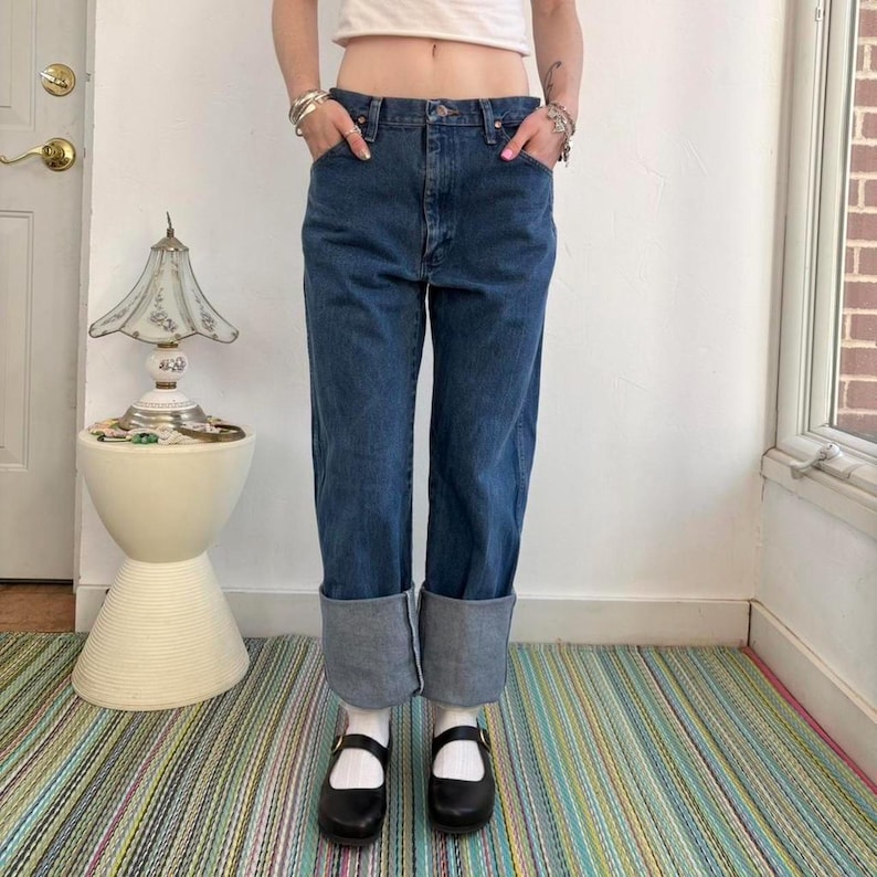 May include: A pair of blue denim jeans with cuffed hems. The jeans have a classic five-pocket design. The model is wearing black Mary Jane shoes with white socks. The jeans are a medium wash denim.