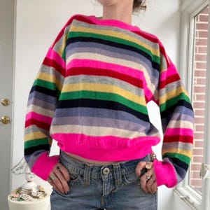 May include: A colorful, striped sweater with a crew neck and long sleeves. The sweater features horizontal stripes in various colors, including pink, red, yellow, green, and blue. The cuffs and hem are a bright pink. The sweater is made of a soft material.