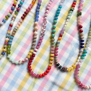 May include: A collection of colorful beaded necklaces in various designs. The necklaces feature a variety of bead shapes and colors, including red, yellow, blue, green, and pink. The necklaces are displayed on a checkered fabric.