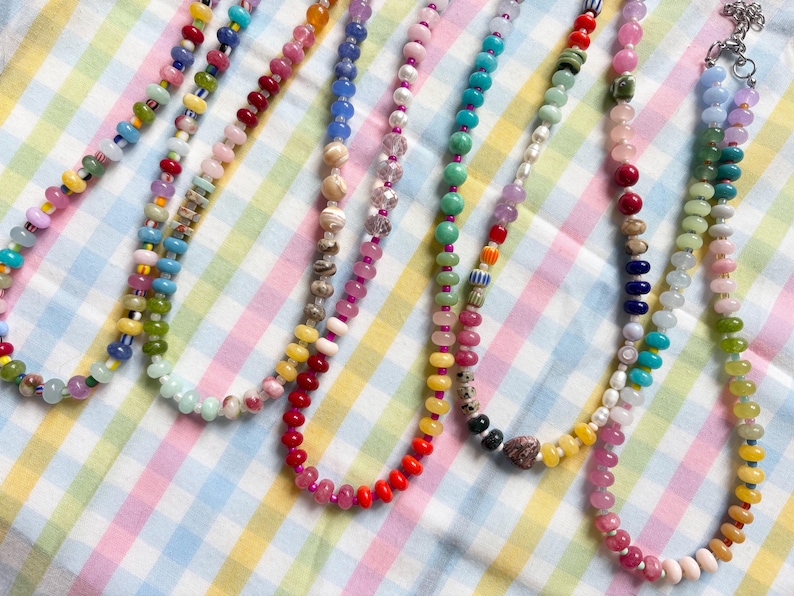 May include: A collection of colorful beaded necklaces in various designs. The necklaces feature a variety of bead shapes and colors, including red, yellow, blue, green, and pink. The necklaces are displayed on a checkered fabric.