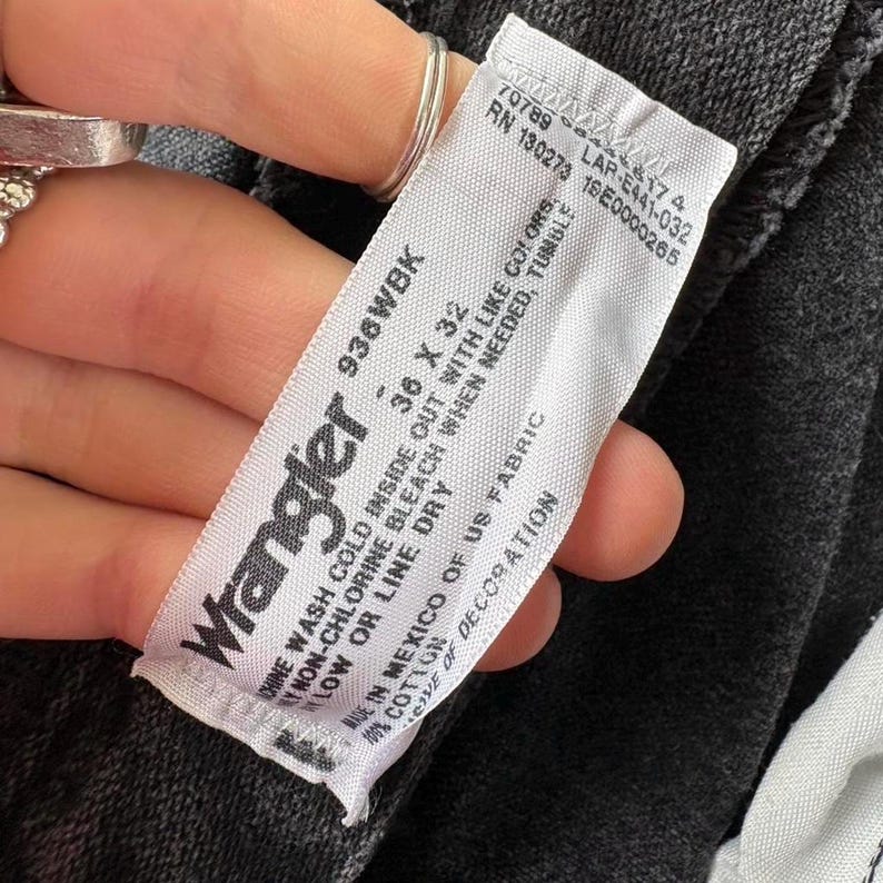 May include: A white Wrangler label with black text, displaying the size 36 x 32. The label is held against dark grey denim jeans. The label also includes washing instructions.