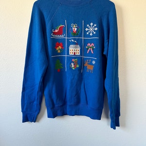 May include: A blue sweatshirt with a Christmas-themed design featuring a grid of nine embroidered images, including a Santa sleigh, a Christmas tree, a house, a reindeer, and a snowflake.
