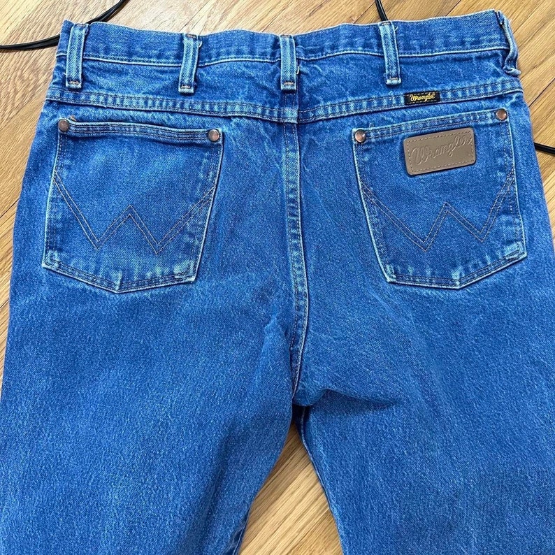 May include: Close-up of the back of blue denim jeans. The jeans have two back pockets with a stitched 'W' design and a brown leather patch with the 'Wrangler' logo. The jeans are on a light brown wooden surface.
