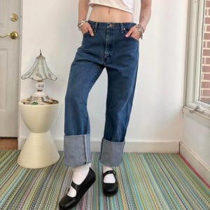 May include: Blue denim jeans with cuffed hems. The jeans have a classic five-pocket design and a straight leg fit. The cuffs are a lighter shade of blue. The person is wearing black Mary Jane shoes and white socks.