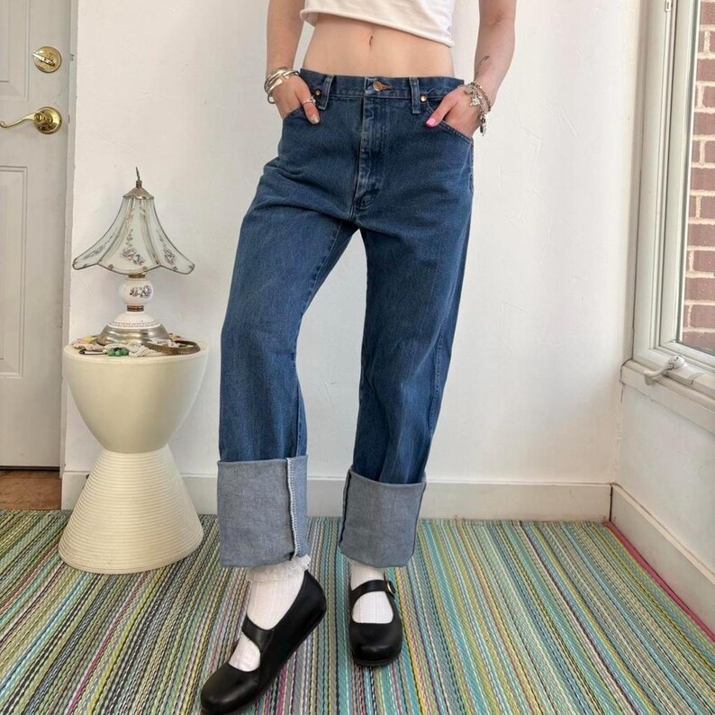 May include: Blue denim jeans with cuffed hems. The jeans have a classic five-pocket design and a straight leg fit. The cuffs are a lighter shade of blue. The person is wearing black Mary Jane shoes and white socks.