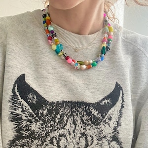 May include: A colorful beaded necklace with a variety of shapes and sizes of beads. The necklace is layered and worn over a gray sweatshirt with a black graphic of a cat's face.