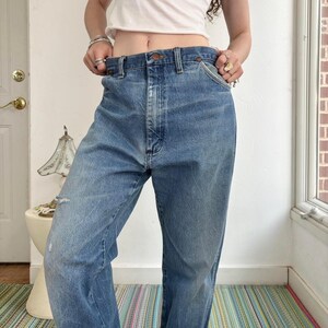 May include: Light blue denim jeans with a classic five-pocket design. The jeans have a straight leg fit and a faded wash, with a distressed area on the left leg. The jeans are worn at the waist.
