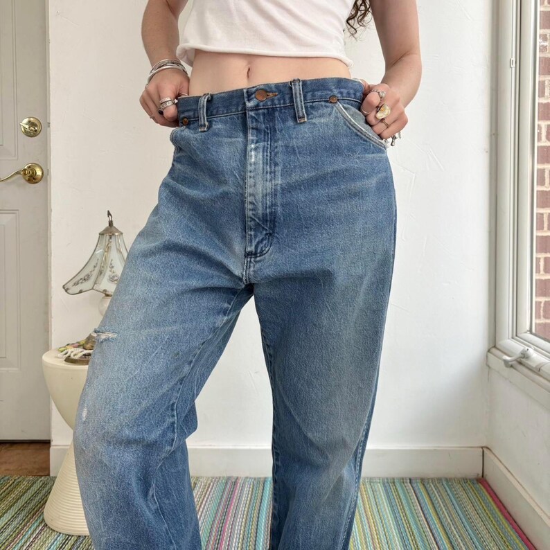 May include: Light blue denim jeans with a classic five-pocket design. The jeans have a straight leg fit and a faded wash, with a distressed area on the left leg. The jeans are worn at the waist.