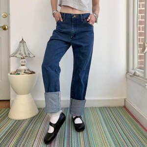 May include: Dark blue denim jeans with cuffed hems. The jeans have a high waist and a classic five-pocket design. The model is wearing black Mary Jane shoes and white socks. The jeans are a vintage style.