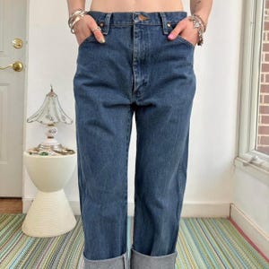 May include: Dark wash denim jeans with a classic five-pocket design. The jeans have a straight leg and are cuffed at the ankles. The model is wearing the jeans with their hands in the pockets. The jeans are a medium-dark blue color.