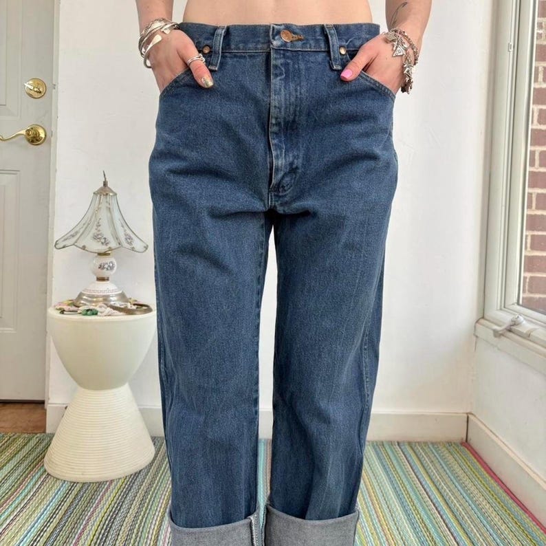 May include: Dark wash denim jeans with a classic five-pocket design. The jeans have a straight leg and are cuffed at the ankles. The model is wearing the jeans with their hands in the pockets. The jeans are a medium-dark blue color.