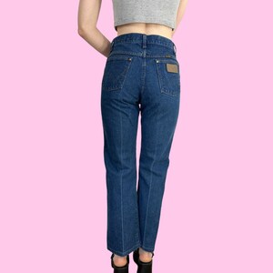 Może przedstawiać: Dżinsy z ciemnego denimu o wysokim stanie i prostym kroju nogawek. Dżinsy mają brązową sk&oacute;rzaną naszywkę z tyłu z logo Wrangler.