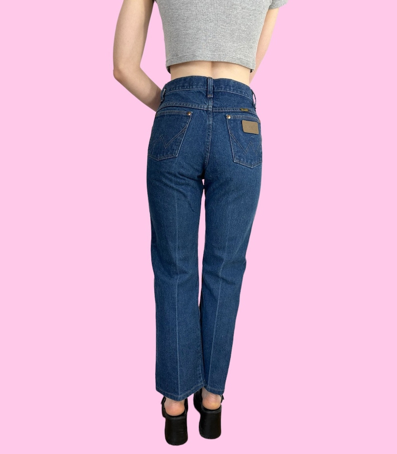 Może przedstawiać: Dżinsy z ciemnego denimu o wysokim stanie i prostym kroju nogawek. Dżinsy mają brązową sk&oacute;rzaną naszywkę z tyłu z logo Wrangler.