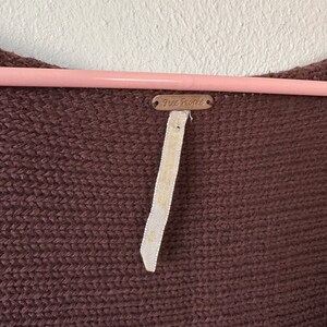 May include: A brown knitted sweater with a Free People tag attached. The tag is a small metal rectangle with the words "Free People" embossed on it.