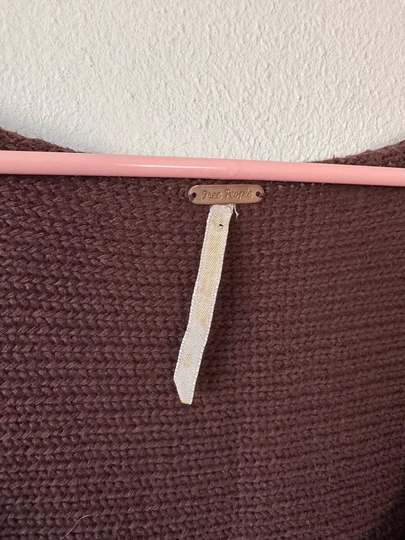 May include: A brown knitted sweater with a Free People tag attached. The tag is a small metal rectangle with the words "Free People" embossed on it.