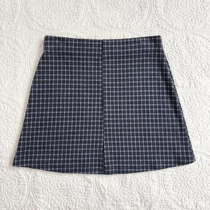 Puede incluir: Falda de corte en A con un patr&oacute;n de cuadros cl&aacute;sico en tonos azul marino, gris y blanco. La falda tiene un dise&ntilde;o sencillo y limpio, con una cintura recta y una ligera forma de l&iacute;nea A. La tela parece ser un material tejido.