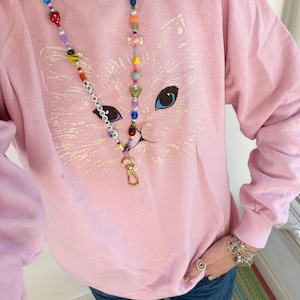May include: A light pink sweatshirt featuring a large, faint white graphic of a cat face with blue eyes. A long lanyard made of colourful, multi-shaped beads hangs around the neck, featuring the text MRS BROOKE and a gold-toned metal lobster clasp.