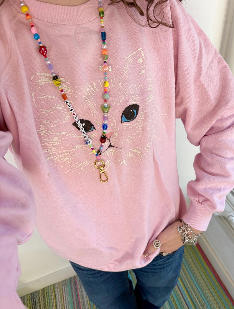 May include: A light pink sweatshirt featuring a large, faint white graphic of a cat face with blue eyes. A long lanyard made of colourful, multi-shaped beads hangs around the neck, featuring the text MRS BROOKE and a gold-toned metal lobster clasp.