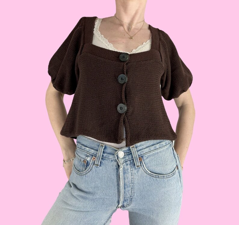 May include: A brown knitted cardigan with puffed sleeves and a square neckline. The cardigan has four black buttons down the front.