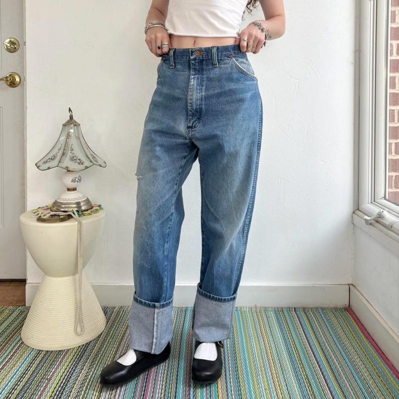 May include: Light blue denim jeans with a high waist and cuffed hems. The jeans have a relaxed fit and a distressed detail on the left knee. The model is wearing black Mary Jane style shoes.