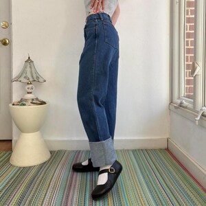 May include: Dark wash denim jeans with a cuffed hem. The jeans are paired with black Mary Jane style shoes with a buckle closure. The person is standing on a striped rug.