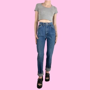 Puede incluir: Un par de pantalones vaqueros azules de mezclilla con cintura alta y corte recto. Los vaqueros tienen un dobladillo con vuelta y se llevan con una camiseta gris recortada y sandalias negras de plataforma.
