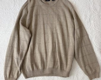 Op de afbeelding: Een beige trui met lange mouwen en een subtiel ruitpatroon op de voorzijde en de mouwen. Het kledingstuk heeft een ronde hals, geribbelde boorden en een geribbelde zoom. Het IZOD-merklogo is zichtbaar in de kraag en als klein geborduurd embleem.