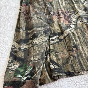 K&ouml;nnte beinhalten: Lang&auml;rmeliges Camouflage-Shirt in Braun-, Gr&uuml;n- und Beiget&ouml;nen. Der Stoff weist ein detailliertes Muster auf, das einer Waldumgebung &auml;hnelt. Der &Auml;rmel des Hemdes ist sichtbar und zeigt die Manschette und die Gesamtstruktur des Materials.