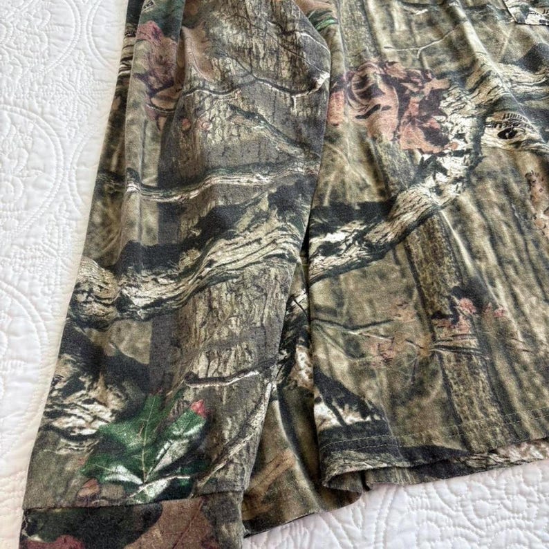 K&ouml;nnte beinhalten: Lang&auml;rmeliges Camouflage-Shirt in Braun-, Gr&uuml;n- und Beiget&ouml;nen. Der Stoff weist ein detailliertes Muster auf, das einer Waldumgebung &auml;hnelt. Der &Auml;rmel des Hemdes ist sichtbar und zeigt die Manschette und die Gesamtstruktur des Materials.