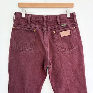 May include: Burgundy-colored Wrangler jeans, displayed on a wooden hanger. The back pockets feature the iconic Wrangler stitching. The jeans have a leather patch and brass rivets. The overall style is classic and casual.