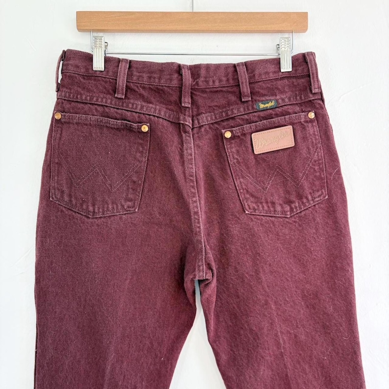 May include: Burgundy-colored Wrangler jeans, displayed on a wooden hanger. The back pockets feature the iconic Wrangler stitching. The jeans have a leather patch and brass rivets. The overall style is classic and casual.