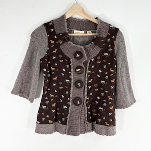 May include: A brown and gray cardigan with a floral print featuring deer, birds, and other woodland creatures. The cardigan has a button closure and a scalloped hem.
