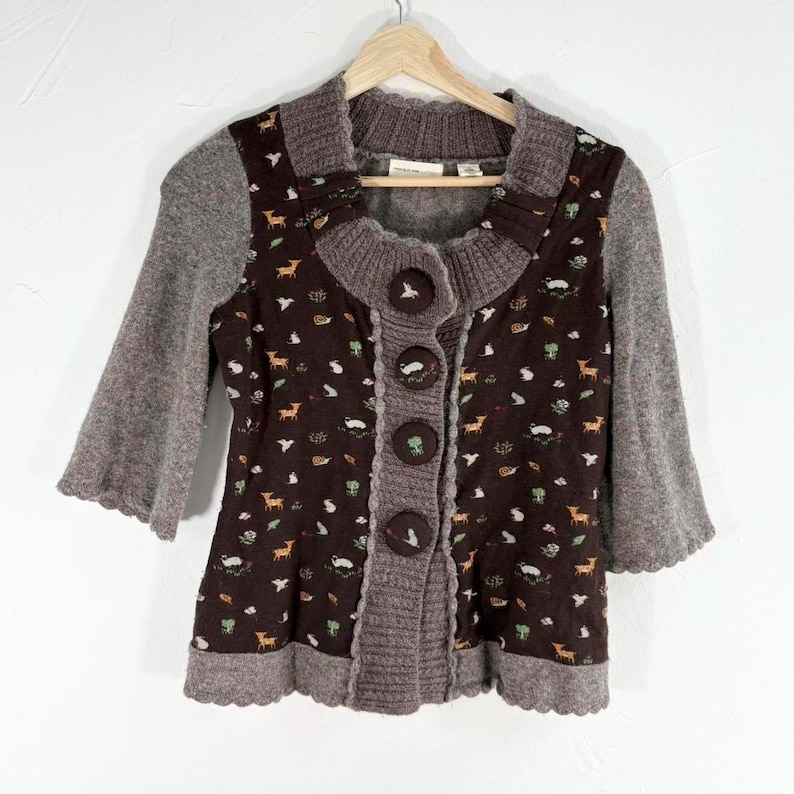May include: A brown and gray cardigan with a floral print featuring deer, birds, and other woodland creatures. The cardigan has a button closure and a scalloped hem.