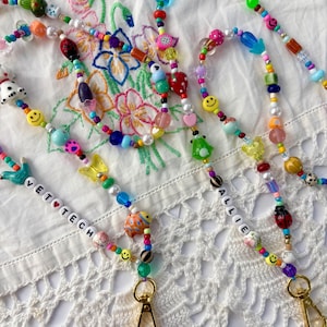 May include: Two beaded lanyards featuring a variety of colourful plastic beads, including smiley faces, butterflies, ladybugs, and fruit shapes. One lanyard features white letter beads spelling VET TECH, and the other spells ALLIE, both with gold clasps.