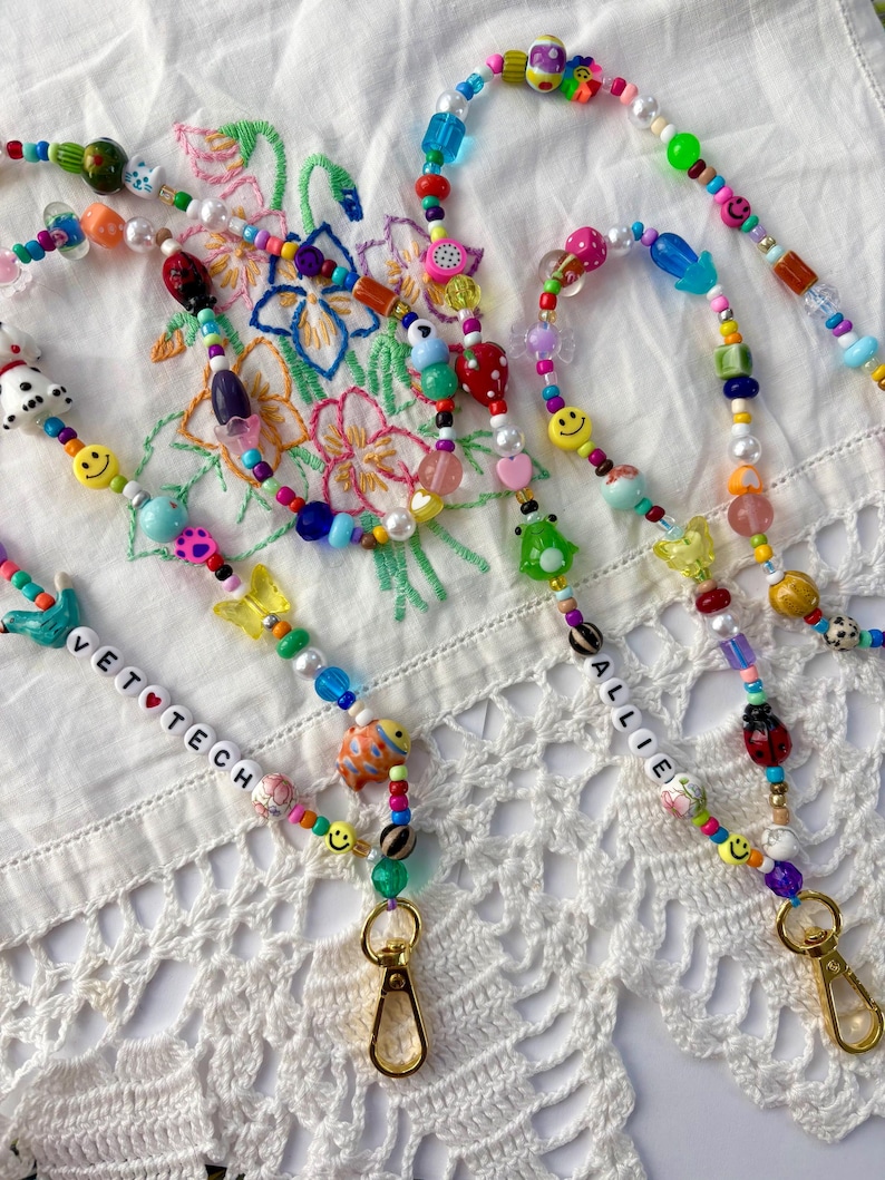 May include: Two beaded lanyards featuring a variety of colourful plastic beads, including smiley faces, butterflies, ladybugs, and fruit shapes. One lanyard features white letter beads spelling VET TECH, and the other spells ALLIE, both with gold clasps.