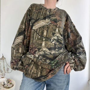 K&ouml;nnte beinhalten: Ein lang&auml;rmeliges Camouflage-Hemd mit Knopfleiste am Ausschnitt. Das Hemd hat ein Muster aus Braun-, Gr&uuml;n- und Beiget&ouml;nen, das einem Wald &auml;hnelt. Die &Auml;rmel sind lang und locker. Das Hemd ist aus einem weichen Material.