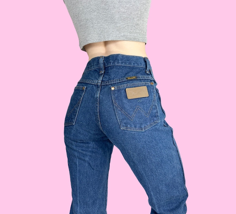 Może przedstawiać: Dżinsy z niebieskiego denimu z brązową sk&oacute;rzaną naszywką na tylnej kieszeni z napisem "Wrangler".