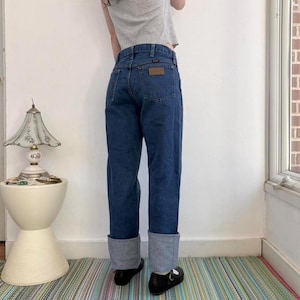 May include: Dark wash denim jeans with a brown leather patch on the back pocket. The jeans have a straight leg fit with a cuffed hem. The person is wearing black shoes.