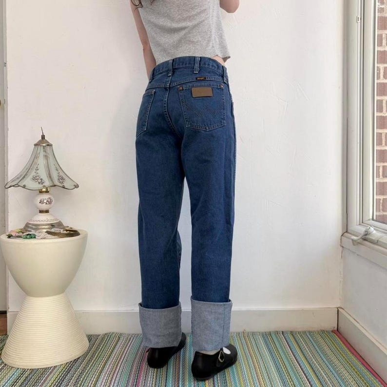 May include: Dark wash denim jeans with a brown leather patch on the back pocket. The jeans have a straight leg fit with a cuffed hem. The person is wearing black shoes.
