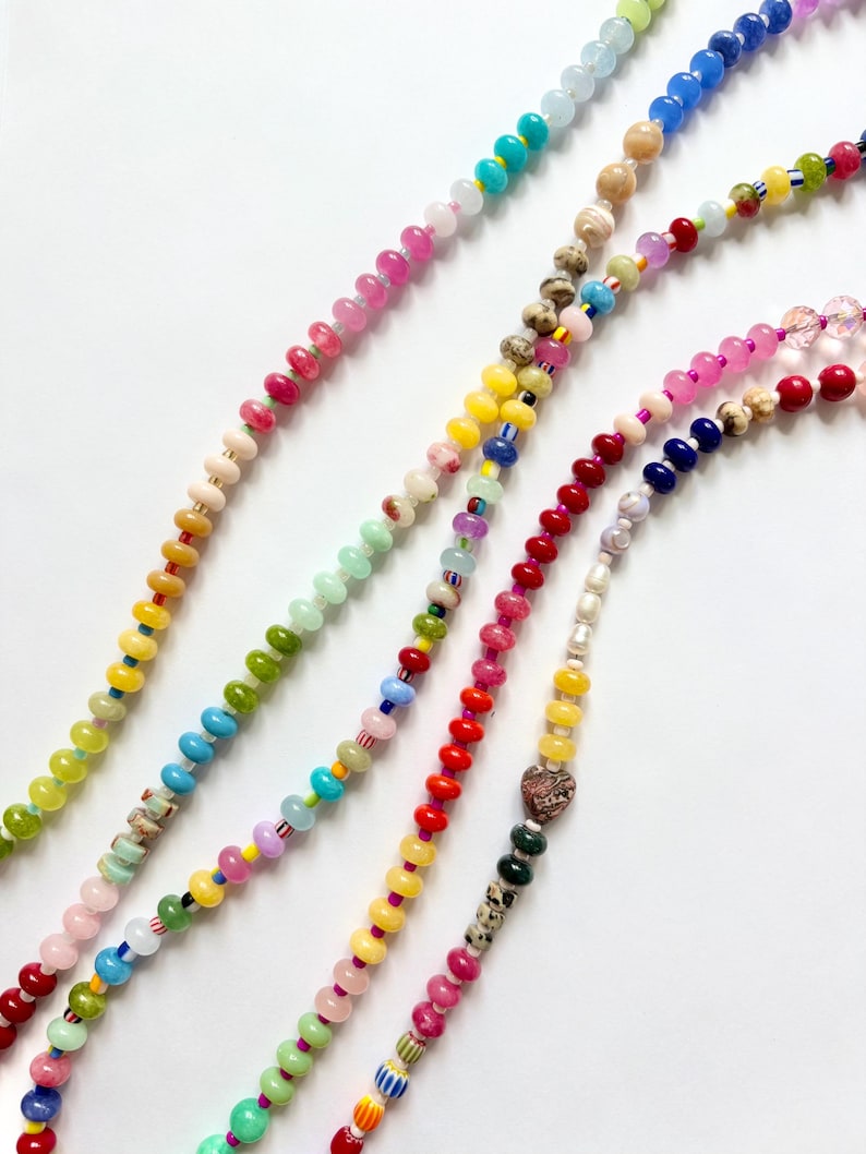 May include: Close-up of several beaded necklaces in various colors, including pink, yellow, green, and blue. The beads are small and round, strung together to create a colorful and eye-catching accessory. The necklaces are displayed against a white background.