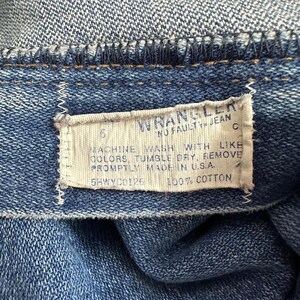 Puede incluir: Una etiqueta de cuidado blanca cosida en una tela vaquera azul. La etiqueta dice "WRANGLER NO FAULT JEAN C 6 MACHINE WASH WITH LIKE COLORS, TUMBLE DRY, REMOVE PROMPTLY MADE IN U.S.A. 5HWYC0126 100% COTTON".
