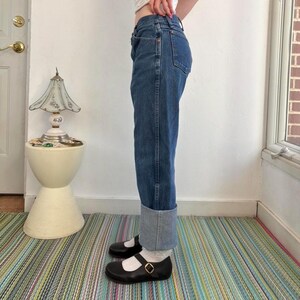 May include: Blue denim jeans with a cuffed hem. The jeans are worn with black Mary Jane style shoes with a gold buckle. The person is standing on a striped rug.