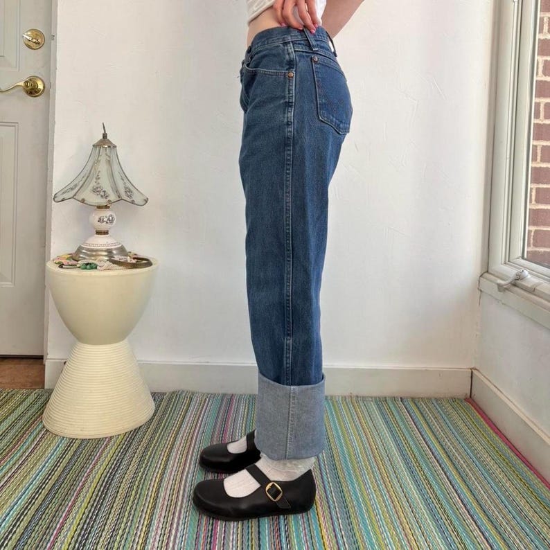 May include: Blue denim jeans with a cuffed hem. The jeans are worn with black Mary Jane style shoes with a gold buckle. The person is standing on a striped rug.
