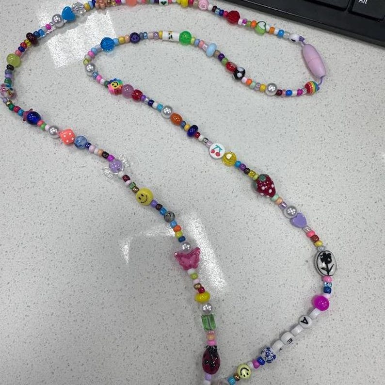 May include: A long beaded lanyard featuring a variety of colourful plastic beads in different shapes and sizes, including smiley faces, a strawberry, a butterfly, a ladybug, cherries, and a flower. The strand includes white letter beads.