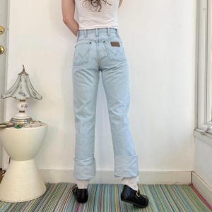 Op de afbeelding: Lichtblauwe jeans met omgeslagen zoom. De jeans heeft een hoge taille en een bruin leren label op de achterzak. De persoon draagt zwarte schoenen. De jeans is in vintage stijl.