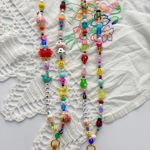 May include: Two beaded lanyards featuring a variety of colourful plastic and glass beads in shapes including animals, fruits, flowers, and smiley faces. One lanyard includes white letter beads spelling VET TECH, and the other spells ALLIE.