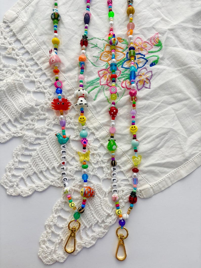 May include: Two beaded lanyards featuring a variety of colourful plastic and glass beads in shapes including animals, fruits, flowers, and smiley faces. One lanyard includes white letter beads spelling VET TECH, and the other spells ALLIE.