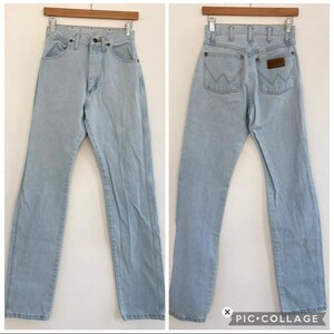 Op de afbeelding: Lichtblauwe denim jeans met een klassiek vijf-zakken ontwerp. De jeans worden van voren en van achteren getoond, met het Wrangler-merklogo zichtbaar op de achterzak. De jeans hangen aan een houten hanger.