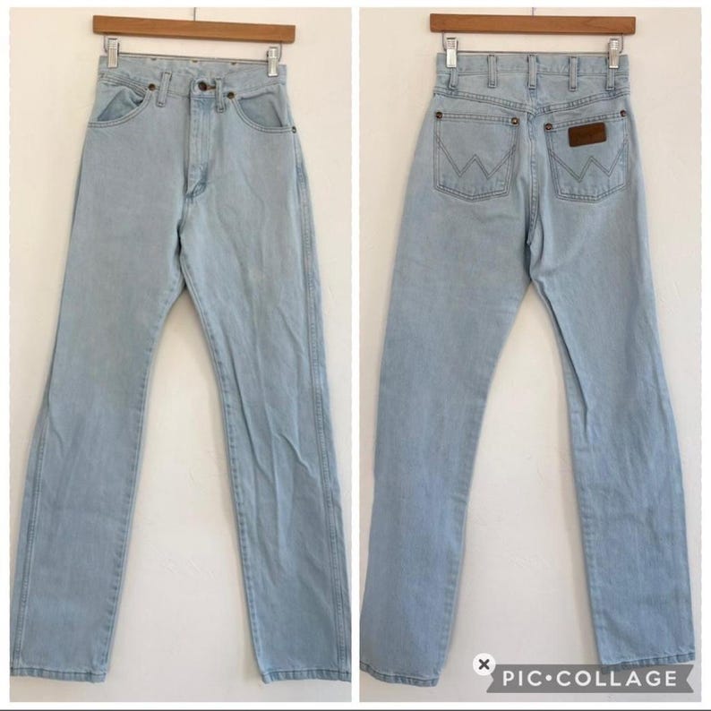 Op de afbeelding: Lichtblauwe denim jeans met een klassiek vijf-zakken ontwerp. De jeans worden van voren en van achteren getoond, met het Wrangler-merklogo zichtbaar op de achterzak. De jeans hangen aan een houten hanger.