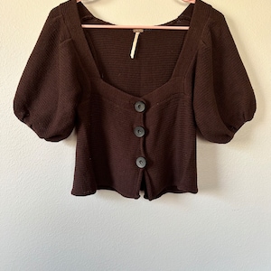 May include: A brown knitted cardigan with a square neckline and puff sleeves. It has a button closure and a cropped length.